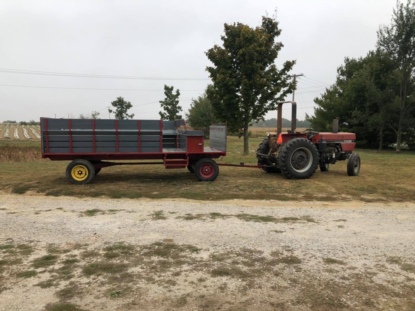 120219153_2938491862916961_863233823773058404_n Red tractor with a trailer parked on a gravel road, surrounded by trees and farmland. The trailer is designed for transporting goods, featuring a flatbed and side rails. In the background, rows of crops are visible under a cloudy sky.
