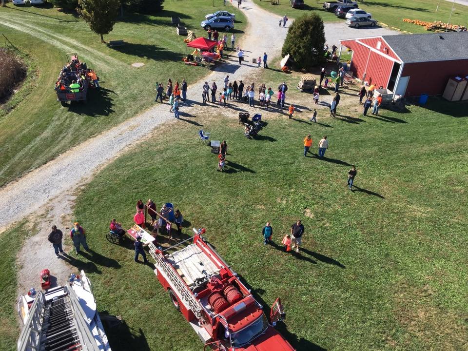 14581396_904870526279115_1307314192823675215_n Aerial view of a busy outdoor event at a farm, featuring families and children participating in activities, a red fire truck, and various attractions including food stalls and a pumpkin patch.