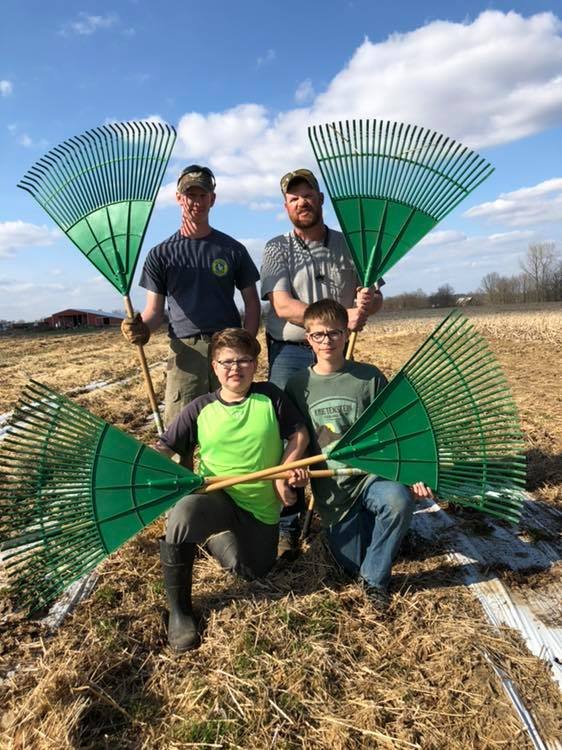 56344478_1837141643051994_7615488009542041600_n Four individuals, including two children, pose outdoors in a field, each holding large green rakes. The background features a clear blue sky and a distant farm structure, illustrating a day of outdoor farming activities.