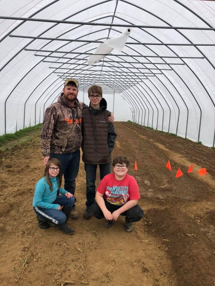 Family of four standing inside a greenhouse, smiling together. The father is wearing a camouflage jacket and cap, while the children are casually dressed. The interior features rows of freshly tilled soil with orange flags marking the planting area, and a white object hanging from the ceiling.